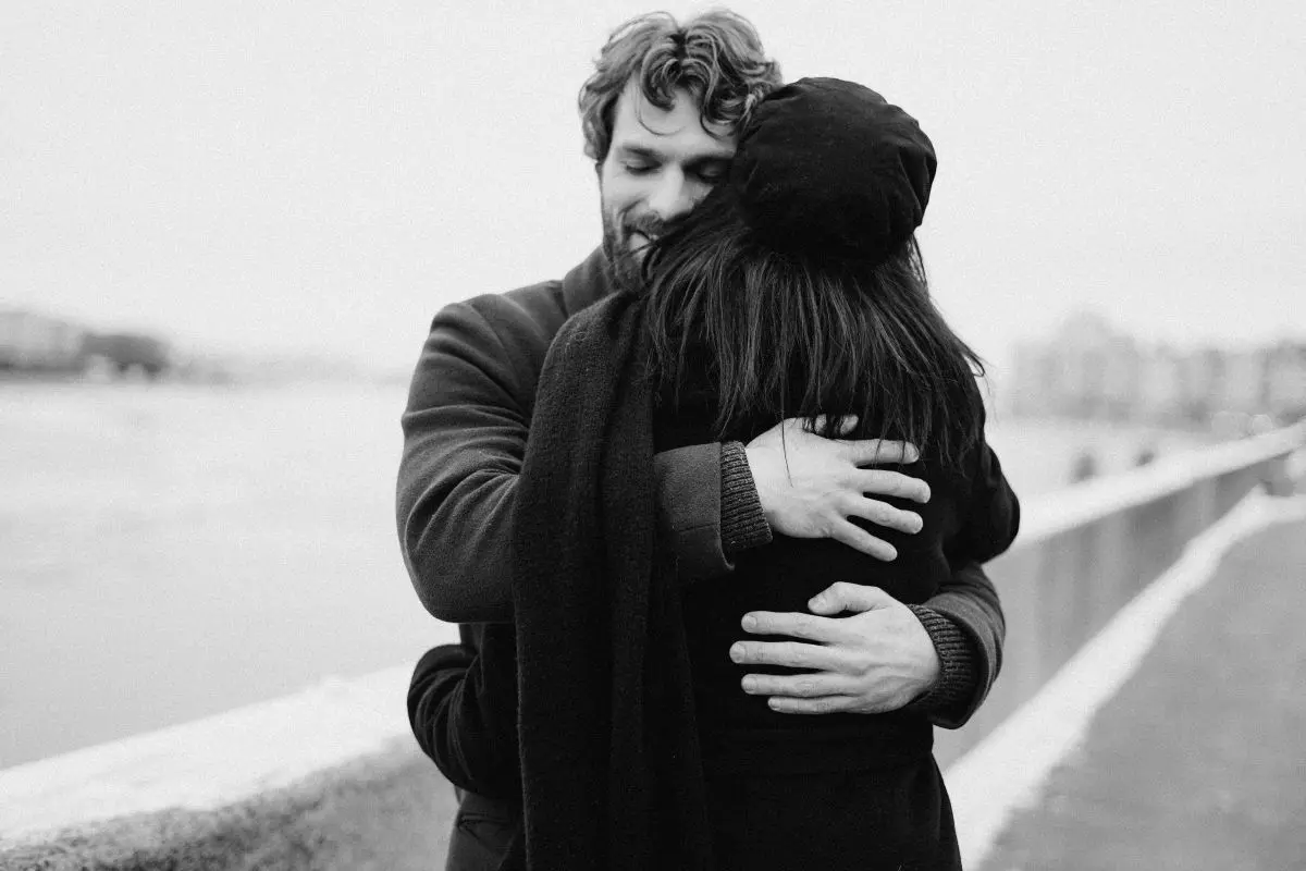 A man with his eyes closed gently hugs a woman in a black beret, photographed in black and white.