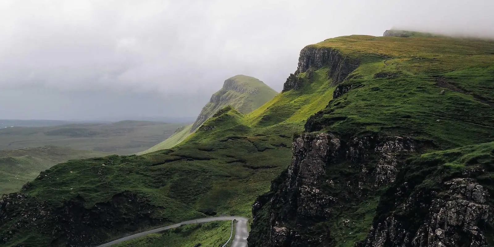 A winding road through a dramatic green mountain range with steep, rocky cliffs under a cloudy sky.