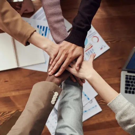 Overhead view of six people with different skin tones stacking their hands on top of each other, symbolizing teamwork. The hands are centered over business documents and a laptop keyboard.
