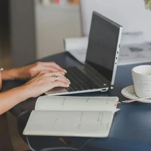 Close-up of woman's hands typing on a silver laptop at a dark blue table, with an open planner/notebook and a white teacup and saucer in the foreground.